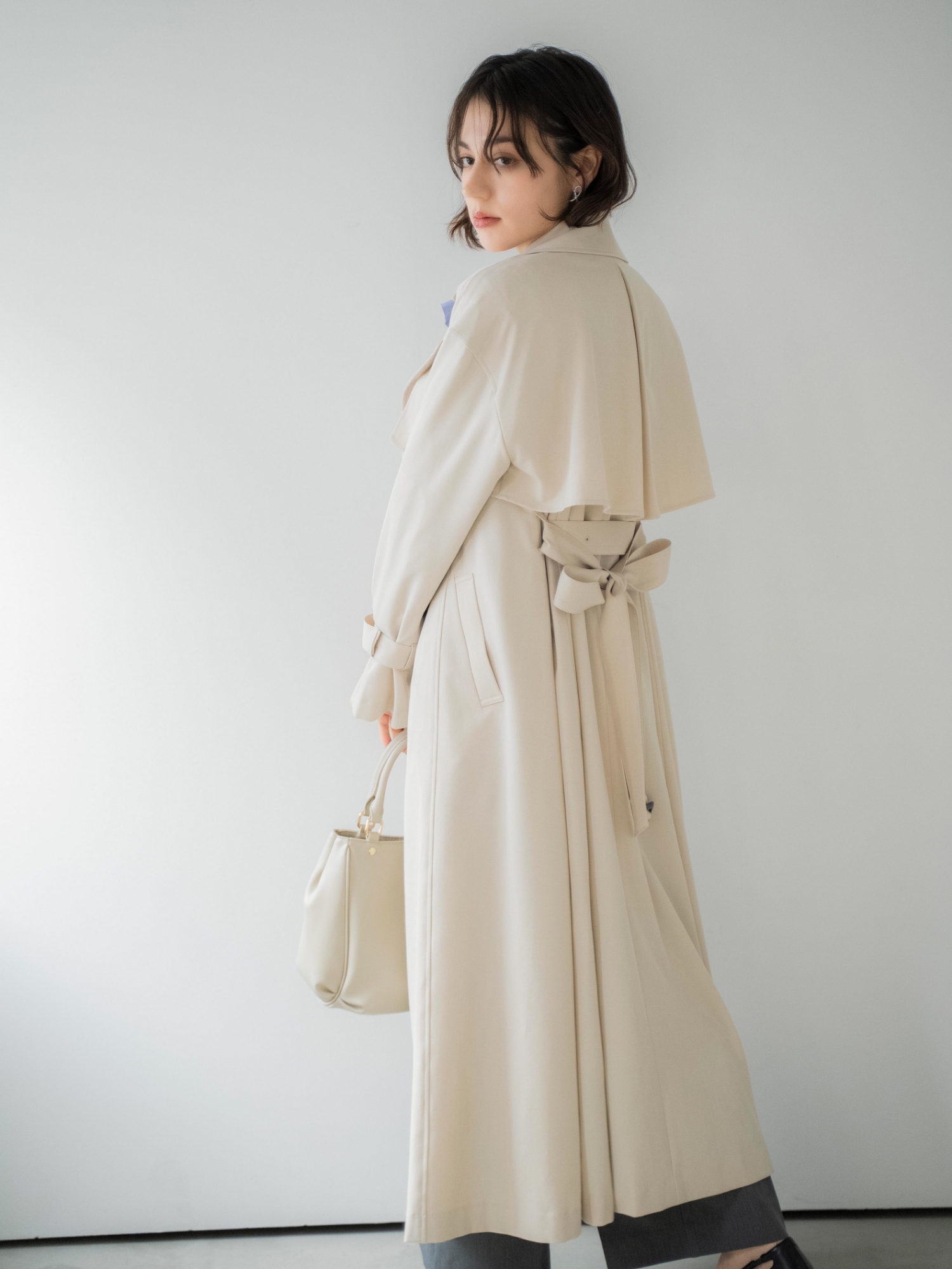 オーバートレンチコート・フレアカラータイブラウス・ポケットデザインワイドパンツ・ツイストデザインパールピアス・ベルトデザインバッグ・サイドノットパンプスを着用した女性の画像