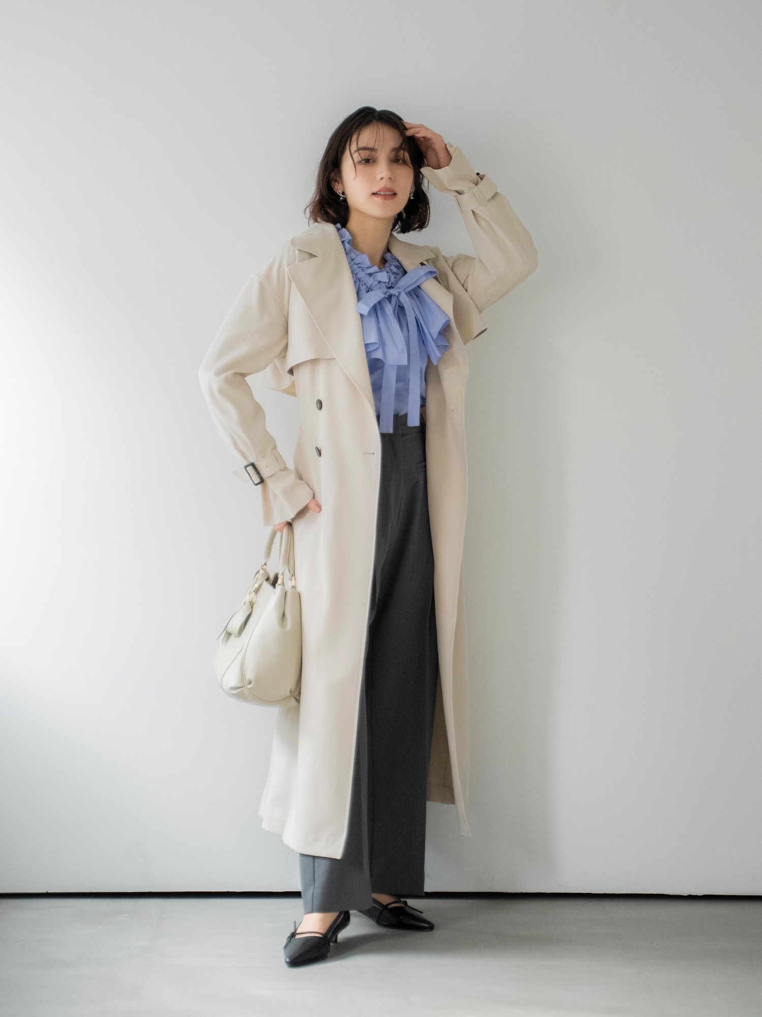 オーバートレンチコート・フレアカラータイブラウス・ポケットデザインワイドパンツ・ツイストデザインパールピアス・ベルトデザインバッグ・サイドノットパンプスを着用した女性の画像